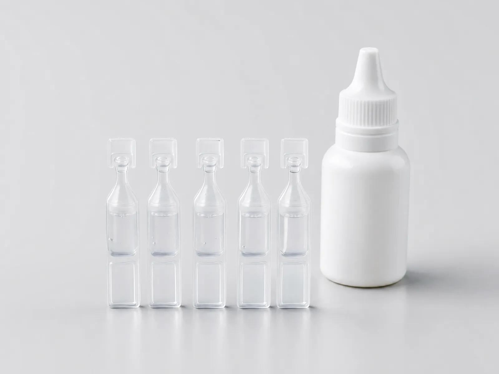 A row of five single-use eye drop vials and one squeeze bottle arranged on a pale grey studio surface