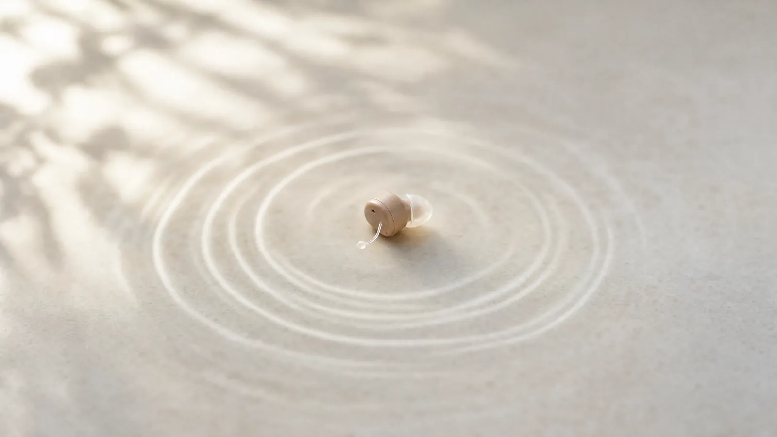 A single hearing aid at the centre of a pale surface surrounded by soft concentric ripples suggesting a persistent ringing tone